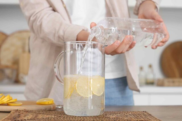 Eine Person gießt frisches Wasser aus einer Plastikflasche in einen Glaskrug, der bereits mit mehreren gelben Zitronenscheiben gefüllt ist. Im Hintergrund ist eine helle Küche angedeutet.