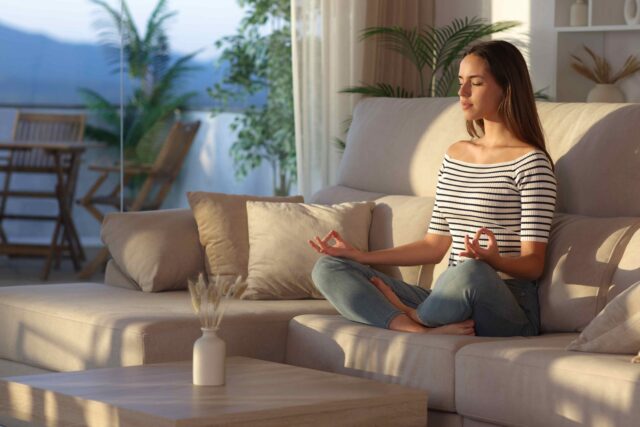 Eine junge Frau meditiert im Schneidersitz auf einem hellen Sofa in einem lichtdurchfluteten Wohnzimmer.