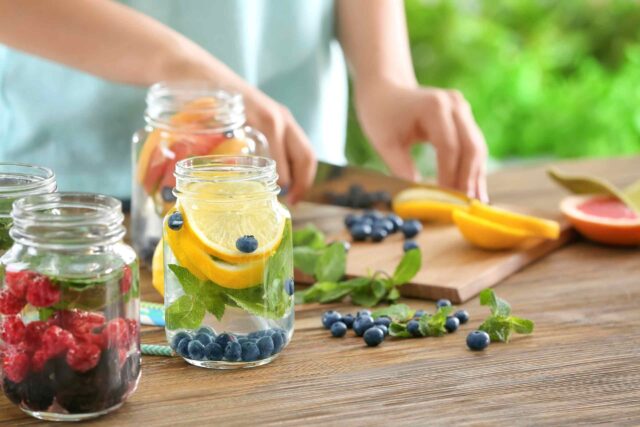 Verschiedene Einmachgläser auf einem Holztisch sind mit Wasser, frischen Beeren, Zitronenscheiben und Minze gefüllt. Im Hintergrund schneidet eine Person weitere Früchte auf einem Holzbrett vor einer grünen Gartenkulisse.