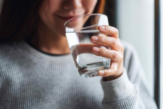 Eine lächelnde Frau hält ein Glas mit klarem Wasser an ihre Lippen, um einen Schluck zu nehmen. Sie trägt einen grauen, strukturierten Pullover; das Licht fällt weich von der Seite ein.