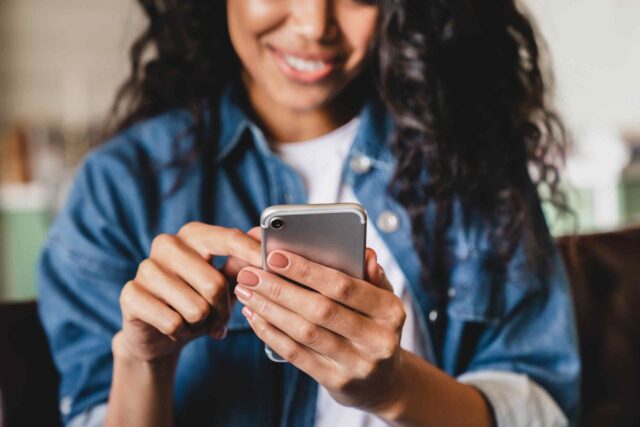 Nahaufnahme einer lächelnden Frau mit dunklem, lockigem Haar, die ein silbernes Smartphone in beiden Händen hält und darauf tippt. Sie trägt eine blaue Jeansjacke über einem weißen T-Shirt.