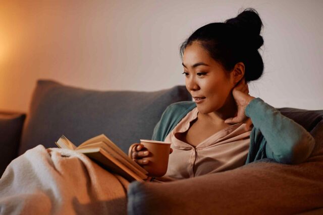 Eine Frau entspannt sich abends auf dem Sofa mit einem Buch und einer Tasse Tee.