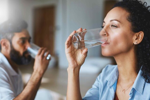 Eine Frau mit dunklen Locken trinkt im Vordergrund aus einem klaren Glas Wasser. Im Hintergrund ist unscharf ein bärtiger Mann zu sehen, der ebenfalls ein Glas Wasser trinkt. Die Szene wirkt hell, sauber und gesundheitsbewusst.