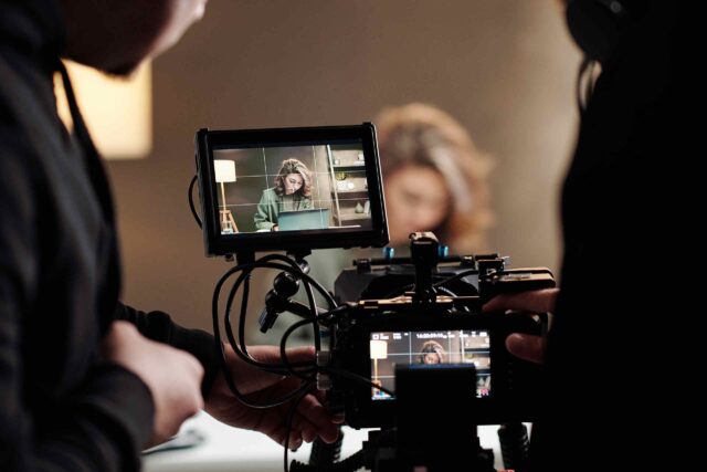 Blick hinter die Kulissen eines Filmdrehs: Im Vordergrund bedienen zwei Personen eine professionelle Filmkamera, deren Monitor eine Frau an einem Laptop zeigt. Die gefilmte Frau sitzt im Hintergrund in einem modern eingerichteten Raum, während das Kamerateam den Bildausschnitt mithilfe von Gitternetzlinien auf dem Display kontrolliert.