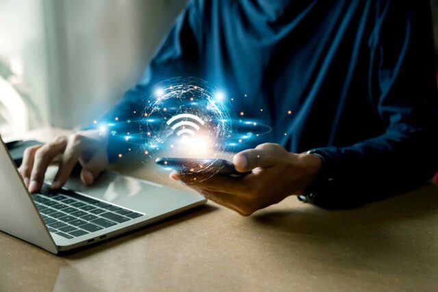 Symbolbild zum Thema moderne Konnektivität: Eine Hand hält ein Smartphone mit einer leuchtenden WLAN-Grafik in einer dunklen Arbeitsumgebung.