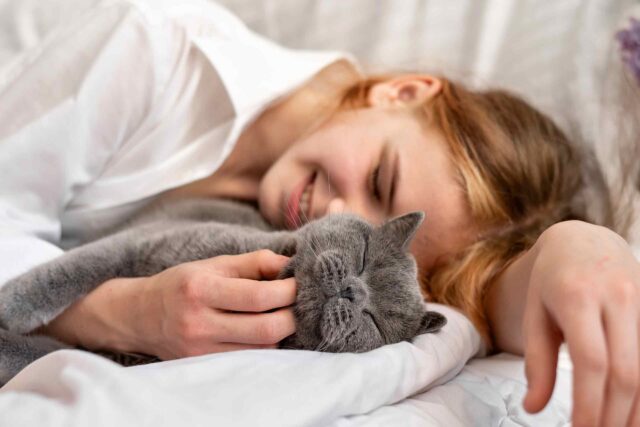 Nahaufnahme einer Frau mit hellbraunem Haar, die lächelnd ihr Gesicht an eine graue British-Kurzhaar-Katze schmiegt. Sie liegen auf weißem Bettzeug, neben ihnen ist ein Strauß Flieder zu sehen.