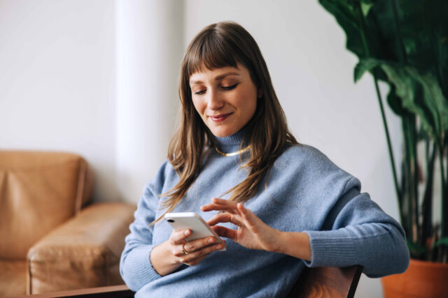Eine lächelnde Frau in einem blauen Rollkragenpullover bedient konzentriert ihr Smartphone.