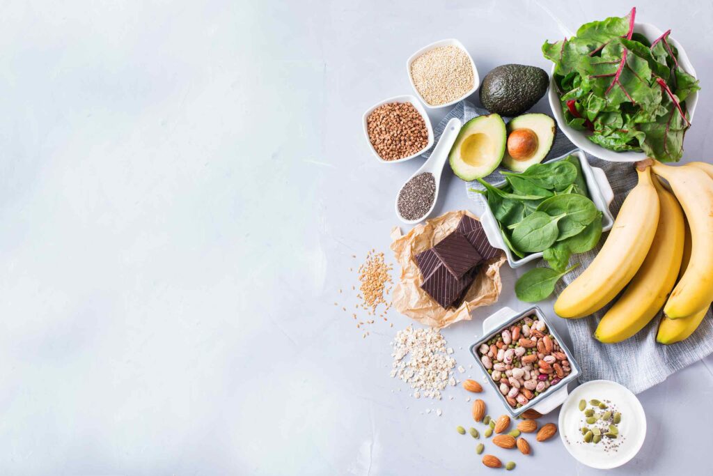 Eine Auswahl an mineralstoffreichen Lebensmitteln wie Bananen, Avocado, dunkle Schokolade, Nüsse, Hülsenfrüchte und grünes Blattgemüse, flach auf hellem Hintergrund arrangiert.