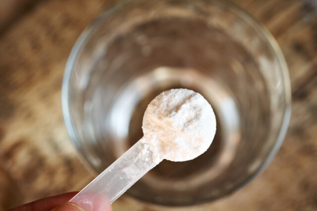 L-Glutamin Pulver in Plastikmessbecher über einem Glas Wasser auf Holztisch
