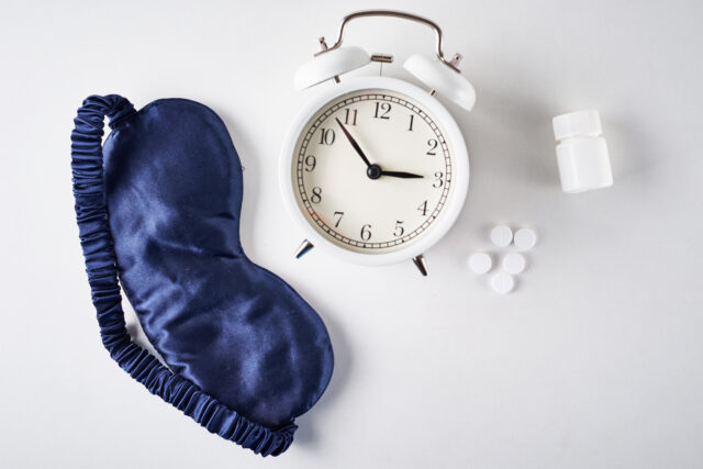 Blaue Schlafmask, weißer Wecker und Melatoninpillen auf weißem Hintergrund