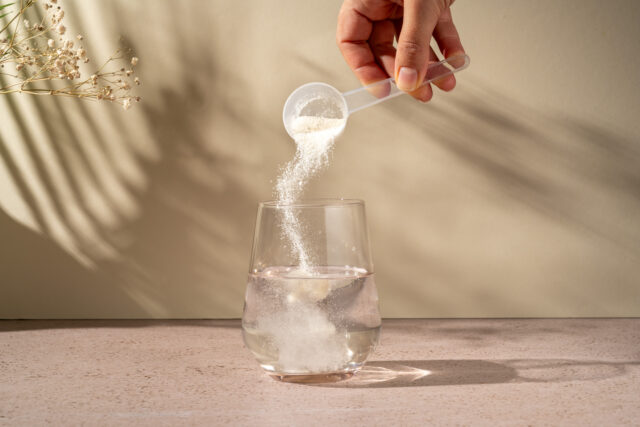 Kollagenpulver im Messlöffel wird in Glas Wasser geschüttet vor beigem Hintergrund