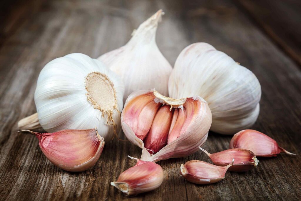 Mehrere ganze Knoblauchknollen und einzelne, rötliche Knoblauchzehen auf einem rustikalen Holztisch.