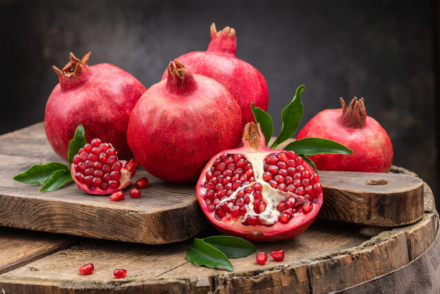 Mehrere Granatäpfel auf einem Tisch vor dunklem Hintergrund, ein Granatapfel ist quer durchgeschnitten.